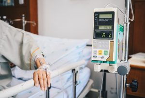 Infusion pump in the foreground of a hospital setting, with intubated patient's hand resting on a bed rail in the background